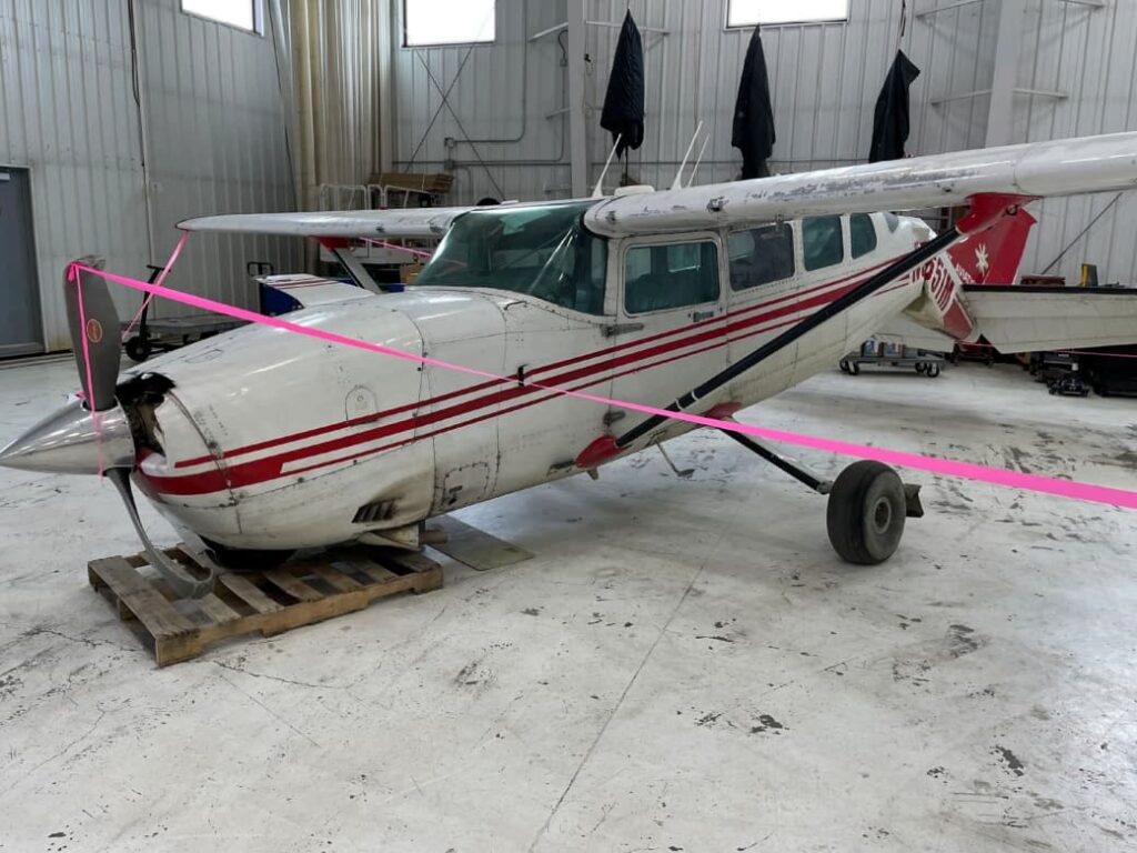 A damaged Cessna 207A aircraft is secured with pink tape inside a maintenance hangar. The aircraft, bearing registration number N9651M and the Grant Aviation logo, shows significant fuselage deformation near the tail section and a bent propeller blade. The airplane is resting on a wooden pallet under the nose, indicating support after a hard landing incident.